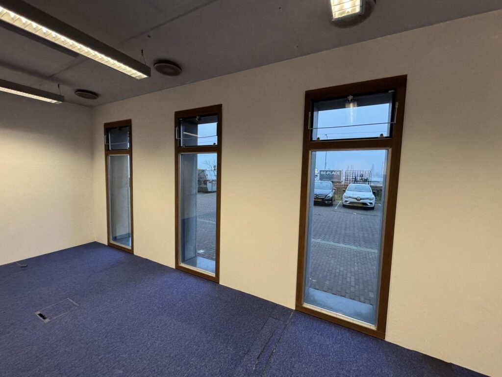 Empty office space with blue carpet, three tall windows, and a view of parked cars outside at De Ruijterkade.